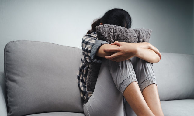 Unhappy lonely sad woman is sitting on the couch and hiding her face on a pillow, depression concept