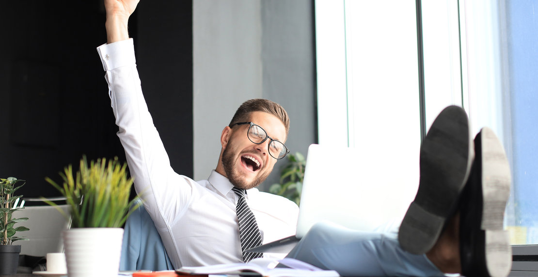 Handsome businessman sitting with legs on table and keeping arm raised and expressing joyful in office